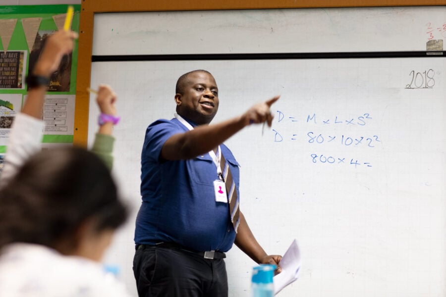 Teacher at the whiteboard pointing to a class of raised hands